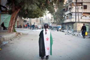 Một bé gái Syria đi trên đường phố của Aleppo, quàng lá cờ 3 sao đối lập với chính quyền Syria (2 sao) trên người. Ảnh chụp hôm 13. 2. 2013 của Bruno Gallado/AFP/Getty